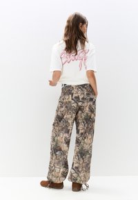 T-shirt blanche avec un texte graphique rose dans le dos, assortie avec un pantalon ample à motif camouflage. Des chaussures marron à lacets viennent parfaire le look.