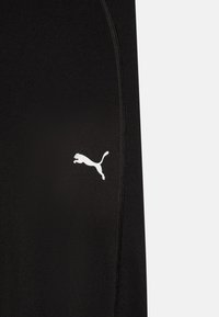 Čierne športové nohavice s hladkou textúrou, s bielym logom Puma a zakriveným švom. Zipsy na bokoch.