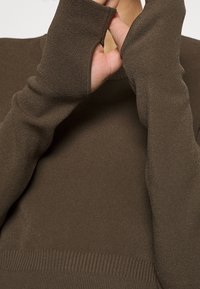 Ruskea neulepaita, jossa pitkät hihat peukalorei'illä, resorireunus ja pehmeä rakenne. Malli on istuva ja minimalistinen.