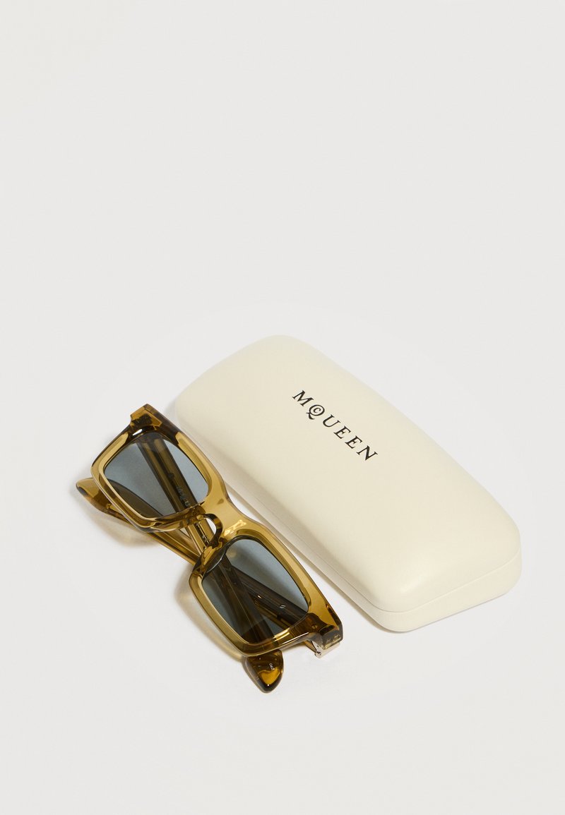 Rectangular amber translucent sunglasses with dark lenses next to a white McQueen branded protective case on a white surface.