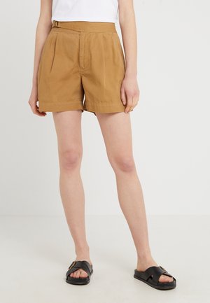 Vrouw draagt hoge bruine korte broek met plooien, een wit topje en zwarte instapsandalen, staand tegen een effen achtergrond.