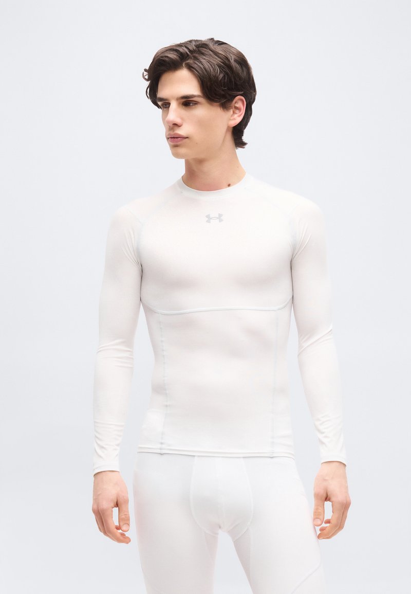 Νέος άνδρας που φοράει εφαρμοστό λευκό αθλητικό μακρυμάνικο τοπ και κολάν, κοιτάζοντας προς τα πλάγια σε απλό φωτεινό φόντο.