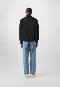 Svart bomberjakke med høy krage, elastiske mansjetter og hem, kombinert med lyseblå løstsittende jeans og hvite joggesko.