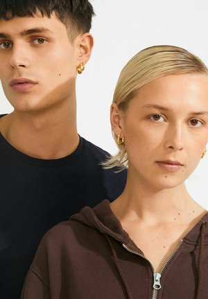 Jonge man met kort donker haar en vrouw met kort blond haar dragen gouden creolen, een donkere sweatshirt en een marineblauw shirt, met neutrale gezichten.