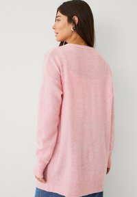 Pull en tricot rose clair avec une coupe décontractée, manches longues et poignets côtelés. Texture douce avec un ourlet arrière légèrement plus long.
