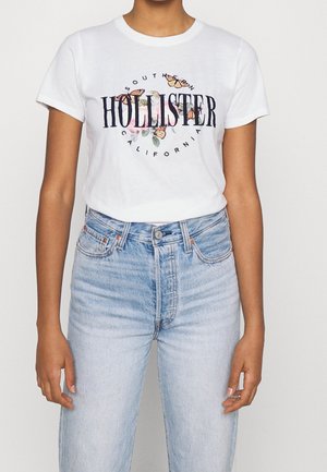 Person iført en hvid Hollister t-shirt med sommerfugl- og blomsterdesign, kombineret med lyseblå højtsiddende jeans.