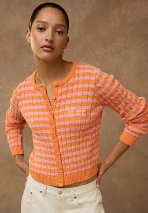 Junge Frau mit dunklem Haar in einem orange- und pink gestreiften, geknöpften Cardigan und hellen Hosen, stehend mit Händen in der Hüfte vor braunem Hintergrund.