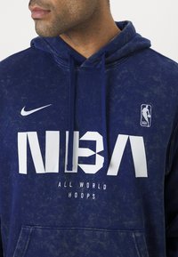 Tamsiai mėlynas džemperis su gobtuvu, turintis tekstūrinį paviršių, su dideliu baltu NBA logotipu ir užrašu "VISAS PASAULIS KREPŠINIS", taip pat su Nike logotipu.