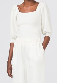 Weißes Top mit Puffärmeln und quadratischem Ausschnitt, aus weichem Stoff. Kombiniert mit cremefarbenen, hoch taillierten, lockeren Hosen.