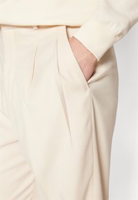 Beżowe spodnie o gładkiej fakturze, z plisowanym wzorem i bocznymi kieszeniami. Pokazane z ręką resting w kieszeni.