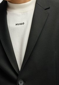 Czarna marynarka o gładkiej fakturze, z pojedynczym guzikiem, nałożona na białą koszulkę z nadrukiem "HUGO" w czerni.