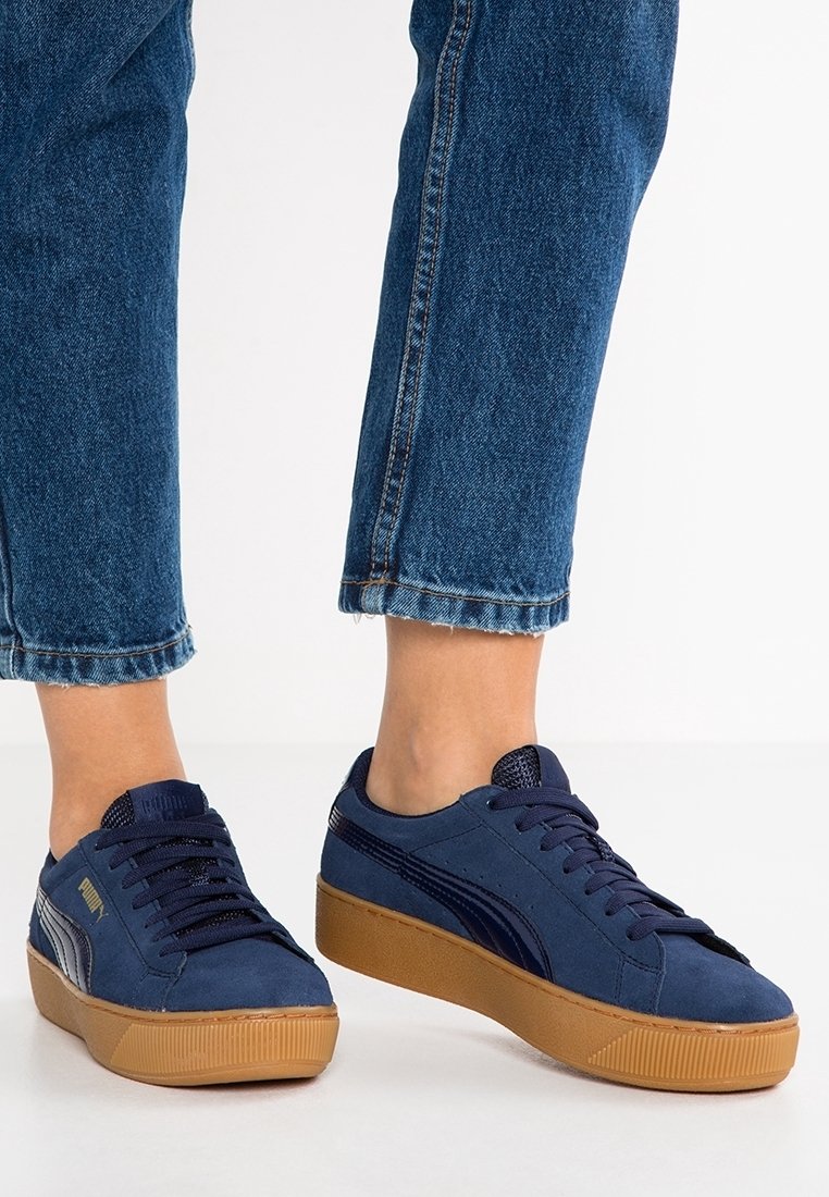 Pieds portant des baskets Puma bleues avec des semelles en gomme et des lacets bleu foncé, associés à un jean en denim bleu raccourci au-dessus des chevilles.
