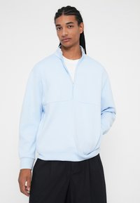 Sweatshirt azul claro com fecho, colarinho alto, textura suave e um contraste branco por baixo. Corte amplo com mangas longas e punhos canelados.