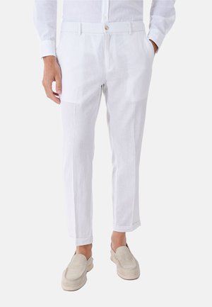 Uomo con pantaloni slim fit bianchi con chiusura a bottone e mocassini beige slip-on, in piedi con le mani in tasca.