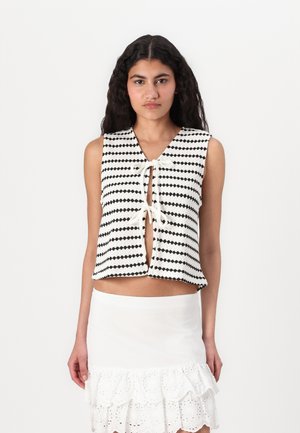 Black and white patterned sleeveless top with a V-neck, tie closure at the front, and cropped design. Paired with a white ruffled skirt.