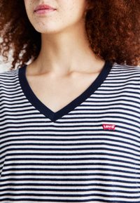 Svītrota kokvilnas T-kreklu ar jūras zila un baltā rakstu; tam ir V-veida kakla izgrieziens un sarkans Levi's logo uzlīme krūtīs.