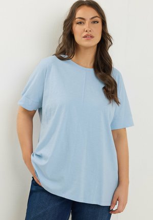 Frau mit langen, welligen braunen Haaren, die ein hellblaues Kurzarmhemd und dunkelblaue Jeans trägt, vor einer weißen Wand stehend.