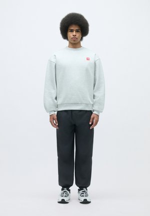 Sudadera gris claro con cuello redondo, puños acanalados y pequeña etiqueta roja; combinada con pantalones oscuros y zapatillas blancas.