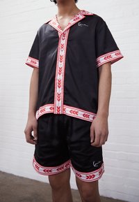 Man wearing black short-sleeve button-up shirt and shorts with red and white leaf-patterned trim, standing indoors against white brick wall.