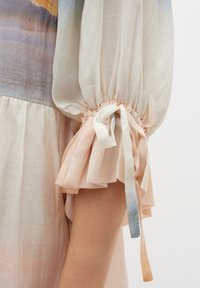 Blouse légère, pastel avec des manches bouffantes. Le tissu texturé est orné de poignets froncés noués avec des accents en ruban.