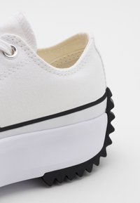 Vita canvas sneakers med en texturerad ovandel, silverögon och en tjock vit sula som har ett svart zigzag-mönster på yttersulan.