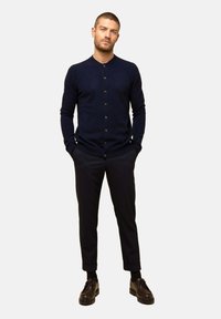 Cárdigan azul marino con botones y tejido texturizado, usado sobre pantalones negros y zapatos negros. La modelo está de pie con las manos en los bolsillos.