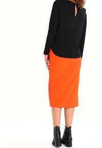 Kvinna iklädd en svart långärmad topp, ljusorange knälång kjol och svarta ankelstövlar, stående vänd bort från kameran mot en vit bakgrund.