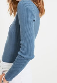 Prążkowana bluzka z długim rękawem w miękkim niebieskim kolorze, o dopasowanym kroju i gładkiej fakturze, zestawiona z białymi spodniami o wysokim stanie.