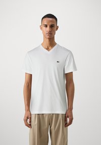 Biely bavlnený tričko s výstrihom do V a krátkymi rukávmi, na ľavej hrudi s malým zeleným logom Lacoste. Hladká textúra, voľný strih.