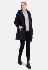 Parka longue marine avec capuche, dotée de poches zippées et d'un léger brillant. Associée à un pull gris et un pantalon noir, complétée par des bottes noires.