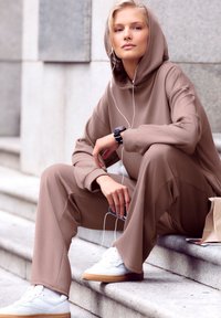 Brown hoodie and matching wide-leg pants made from soft material, paired with white sneakers and headphones, seated on stone steps.