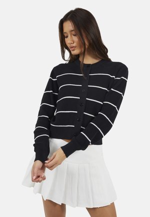 Giovane donna che aggiusta le maniche, indossando un cardigan nero con bottoni e righe bianche e una gonna plissé bianca, su uno sfondo semplice.