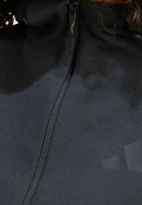 Svart zip-up huvtröja med en mjuk yta, hög krage och subtila logodetaljer. Tyget verkar lätt och flexibelt.