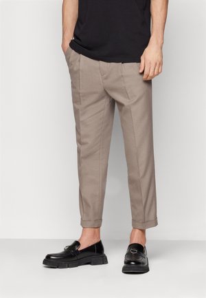 Mężczyzna w beżowych, dopasowanych spodniach z mankietami, czarnych mokasynach i czarnej koszuli, stojący z częściowo wsuniętymi rękami do kieszeni.