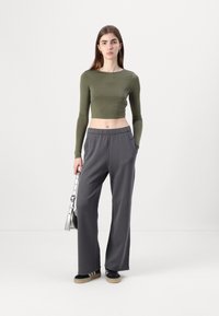 Long-sleeve, olive green crop top paired with loose grey trousers. Model holds a metallic handbag, wearing black sneakers with white stripes.