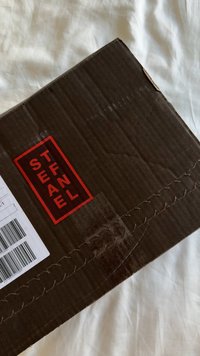 Caja de cartón negro con texto rojo "STEAL THE FINAL" y una etiqueta de envío con códigos de barras, sobre una tela blanca.