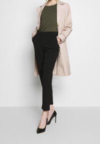 Beige trenčkot, olivově zelené tričko, černé zúžené kalhoty a černé vysoké podpatky. Trenčkot má velké klopy a knoflíky, kalhoty mají boční kapsy.