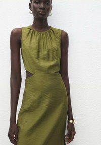 Oliwkowa sukienka bez rękawów z zebranym dekoltem, bocznymi wycięciami i dopasowanym krojem. Gładki materiał o subtelnej fakturze. Złota bransoletka jako akcent.