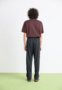 Person med krøllet hår iført en brun t-shirt, sorte bukser og sorte sko med røde accenter, der står på et grønt gulv og ser mod en hvid væg.