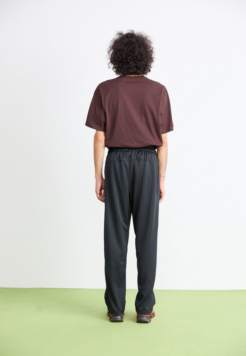 Persona con capelli ricci che indossa una maglietta marrone, pantaloni neri e scarpe nere con accenti rossi, in piedi su un pavimento verde di fronte a un muro bianco.