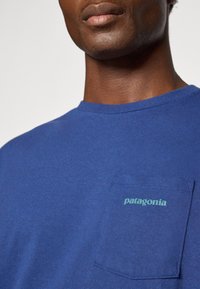 Blauwe katoenen t-shirt met ronde hals en korte mouwen. Kenmerkt zich door een linkse borstzak met het "patagonia" logo in het groen.