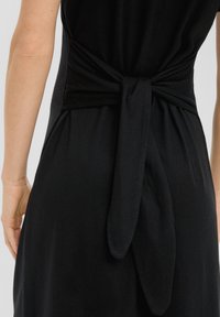 Robe noire avec un détail en nœud à l'arrière, fabriquée en tissu doux. Coupé droit avec une texture lisse et peu d'embellissements.