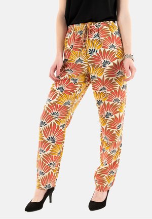 Femme portant un pantalon taille haute avec un motif floral jaune, orange et noir, un haut noir, des talons noirs et une montre-bracelet argentée, debout.