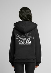 Fekete kapucnis pulóver, nagy fehér grafikával a hátsó részén, rajta a "Lost Treasures" és a "Pequs Művészeti Galéria Múzeum, Mykonos" felirattal.