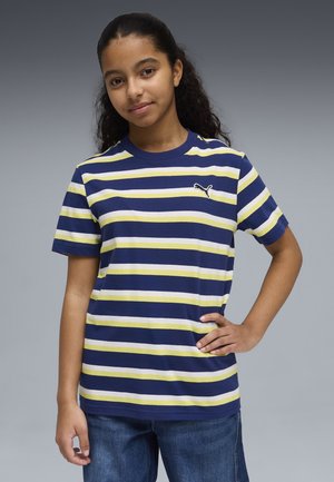 Persona joven con cabello largo y rizado, lleva una camiseta de rayas azul marino, blanca y amarilla de Puma y jeans azules, posando con una mano en la cadera.