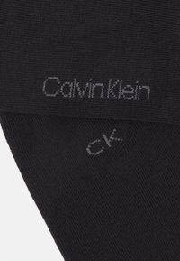 Calcetines negros con textura acanalada que presentan logotipos grises: "Calvin Klein" y "CK". Diseño simple, adecuado para uso casual.