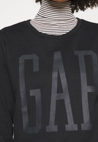 Sudadera negra con gran logo 'GAP' mate en la parte delantera. Presenta un escote redondo y una camiseta de cuello alto a rayas negras y blancas debajo.