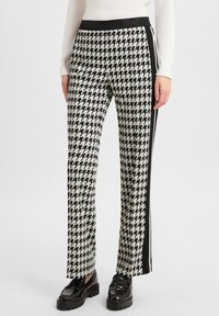 Woman wearing black and white houndstooth pants with black side stripes and black loafers, standing against a plain background.