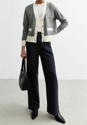 Femme portant un cardigan gris et blanc boutonné, un top blanc, un pantalon large à fines rayures noires, des bottes pointues noires, tenant un sac à main noir.