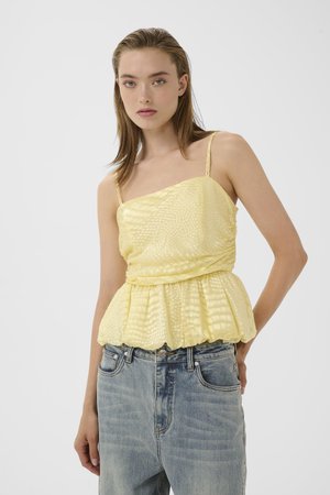 Jeune femme aux cheveux raides portant un top sans manches texturé jaune et un jean en denim bleu clair sur un fond uni.